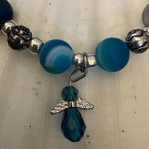 May include: A beaded bracelet featuring blue and silver beads. The bracelet has a dangling charm of a teal angel with silver wings. The beads are various sizes and shapes, with a mix of textures.