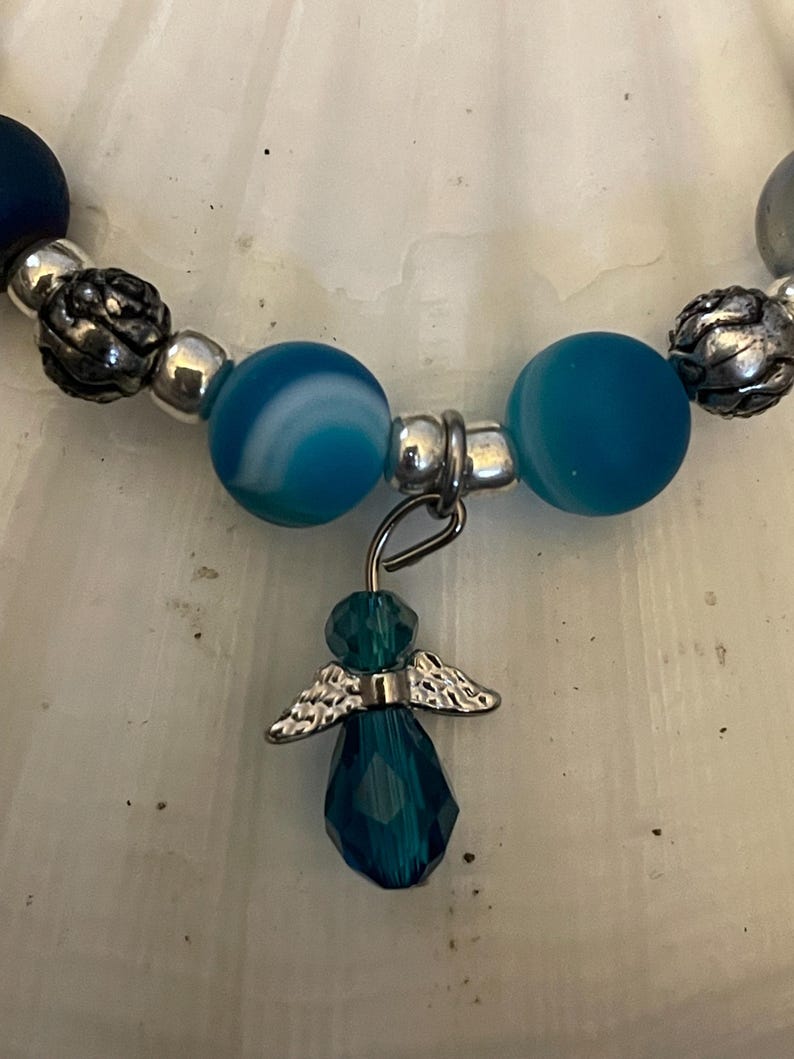 May include: A beaded bracelet featuring blue and silver beads. The bracelet has a dangling charm of a teal angel with silver wings. The beads are various sizes and shapes, with a mix of textures.