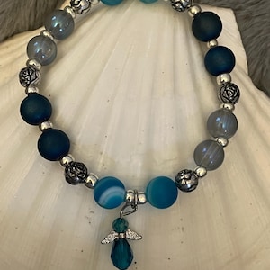 May include: A beaded bracelet featuring a mix of blue and gray beads, interspersed with silver-colored accents. A small angel charm with a teal-colored teardrop pendant hangs from the bracelet. The bracelet is displayed on a white seashell.