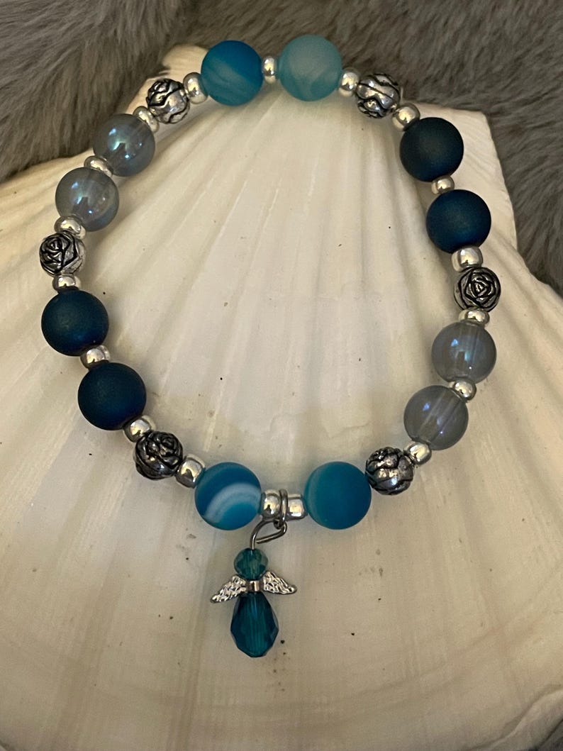 May include: A beaded bracelet featuring a mix of blue and gray beads, interspersed with silver-colored accents. A small angel charm with a teal-colored teardrop pendant hangs from the bracelet. The bracelet is displayed on a white seashell.