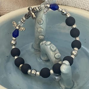 May include: A beaded bracelet featuring black matte beads, silver-toned metal beads, and decorative angel charms. The bracelet also includes blue faceted beads and a small cross charm. The bracelet is displayed on a light blue ceramic surface.