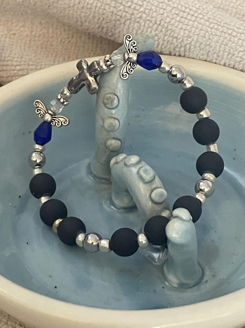 May include: A beaded bracelet featuring black matte beads, silver-toned metal beads, and decorative angel charms. The bracelet also includes blue faceted beads and a small cross charm. The bracelet is displayed on a light blue ceramic surface.