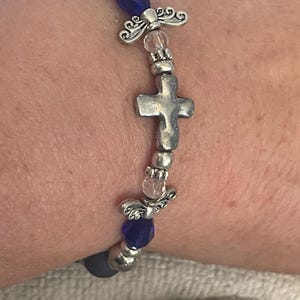 May include: A beaded bracelet featuring a silver cross charm, angel wing accents, and faceted blue beads. The bracelet is strung on a dark blue elastic cord and has silver-toned metal beads.