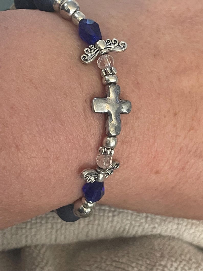 May include: A beaded bracelet featuring a silver cross charm, angel wing accents, and faceted blue beads. The bracelet is strung on a dark blue elastic cord and has silver-toned metal beads.