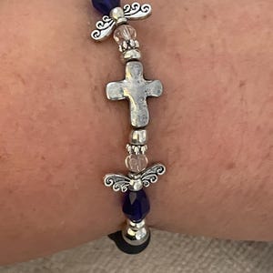 May include: A silver-toned bracelet featuring a cross and angel wing charms, interspersed with blue and clear beads. The bracelet is strung on a black elastic band, creating a religious or spiritual theme.