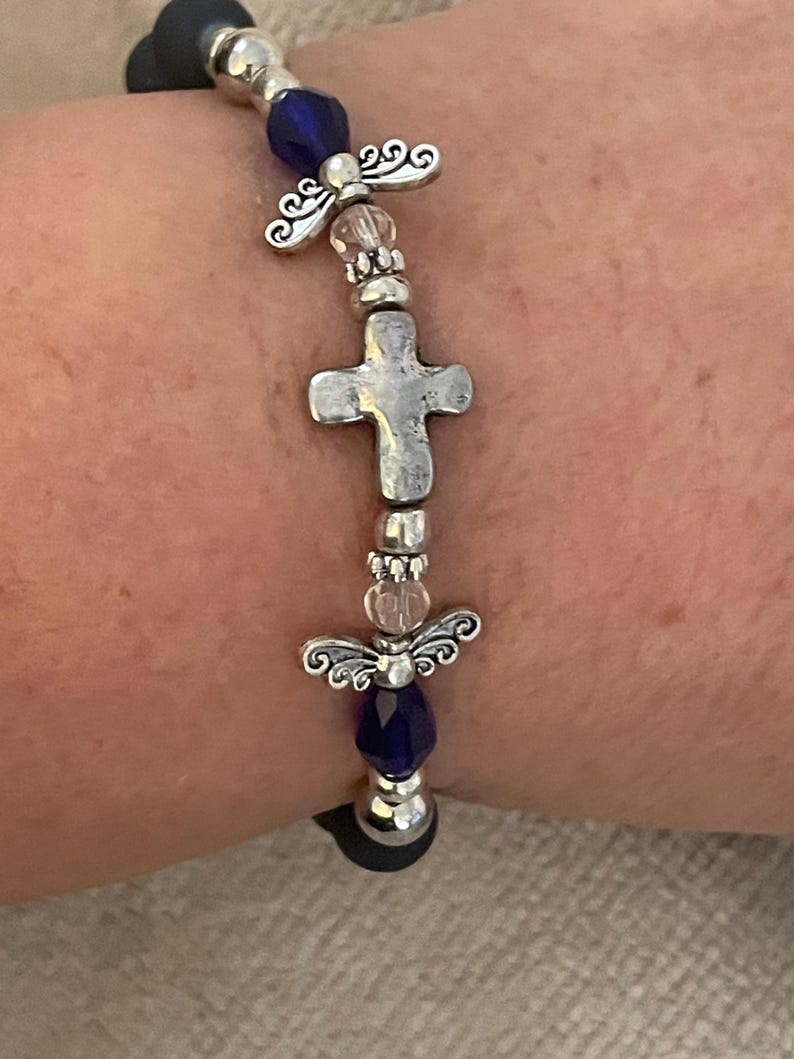 May include: A silver-toned bracelet featuring a cross and angel wing charms, interspersed with blue and clear beads. The bracelet is strung on a black elastic band, creating a religious or spiritual theme.