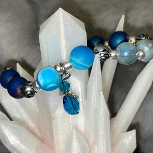 May include: A beaded bracelet featuring various shades of blue beads, including marbled, iridescent, and dark blue, interspersed with silver-colored metal accents. A small, dangling charm with a blue crystal adds a touch of elegance. The bracelet is displayed on a white, crystalline structure.