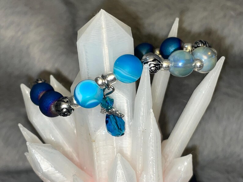 May include: A beaded bracelet featuring various shades of blue beads, including marbled, iridescent, and dark blue, interspersed with silver-colored metal accents. A small, dangling charm with a blue crystal adds a touch of elegance. The bracelet is displayed on a white, crystalline structure.