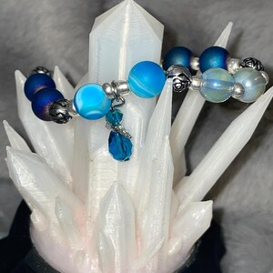 May include: A beaded bracelet featuring blue and silver beads. The bracelet has a mix of matte and shiny beads in shades of blue, with silver accents. A blue crystal charm hangs from the bracelet. The bracelet is displayed on a white crystal.