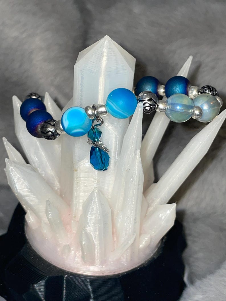May include: A beaded bracelet featuring blue and silver beads. The bracelet has a mix of matte and shiny beads in shades of blue, with silver accents. A blue crystal charm hangs from the bracelet. The bracelet is displayed on a white crystal.