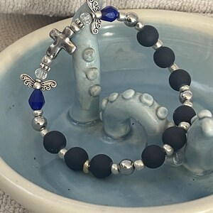 May include: A beaded bracelet featuring matte black beads, silver-toned metal beads, and decorative angel charms. The bracelet includes a silver cross charm and blue accents. The bracelet is displayed on a light blue ceramic dish.
