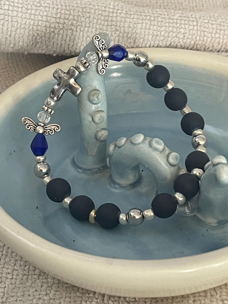 May include: A beaded bracelet featuring matte black beads, silver-toned metal beads, and decorative angel charms. The bracelet includes a silver cross charm and blue accents. The bracelet is displayed on a light blue ceramic dish.