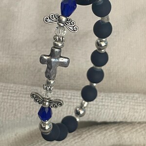 May include: A beaded bracelet featuring dark blue matte beads and silver-toned accents. The bracelet includes a silver cross charm, angel wing charms, and faceted blue beads. The bracelet is designed with a stretchable band.
