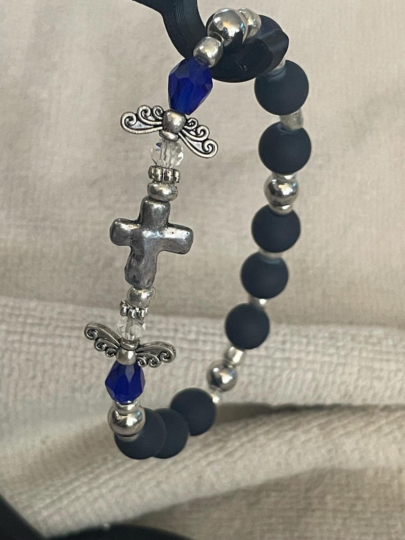 May include: A beaded bracelet featuring dark blue matte beads and silver-toned accents. The bracelet includes a silver cross charm, angel wing charms, and faceted blue beads. The bracelet is designed with a stretchable band.
