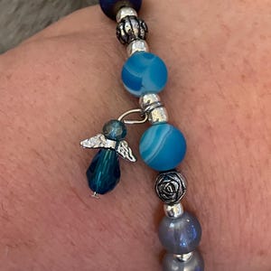 May include: A beaded bracelet featuring blue and silver beads, with a dangling angel charm. The bracelet includes various shades of blue beads, silver accents, and a small angel pendant. The bracelet is designed to be worn on the wrist.