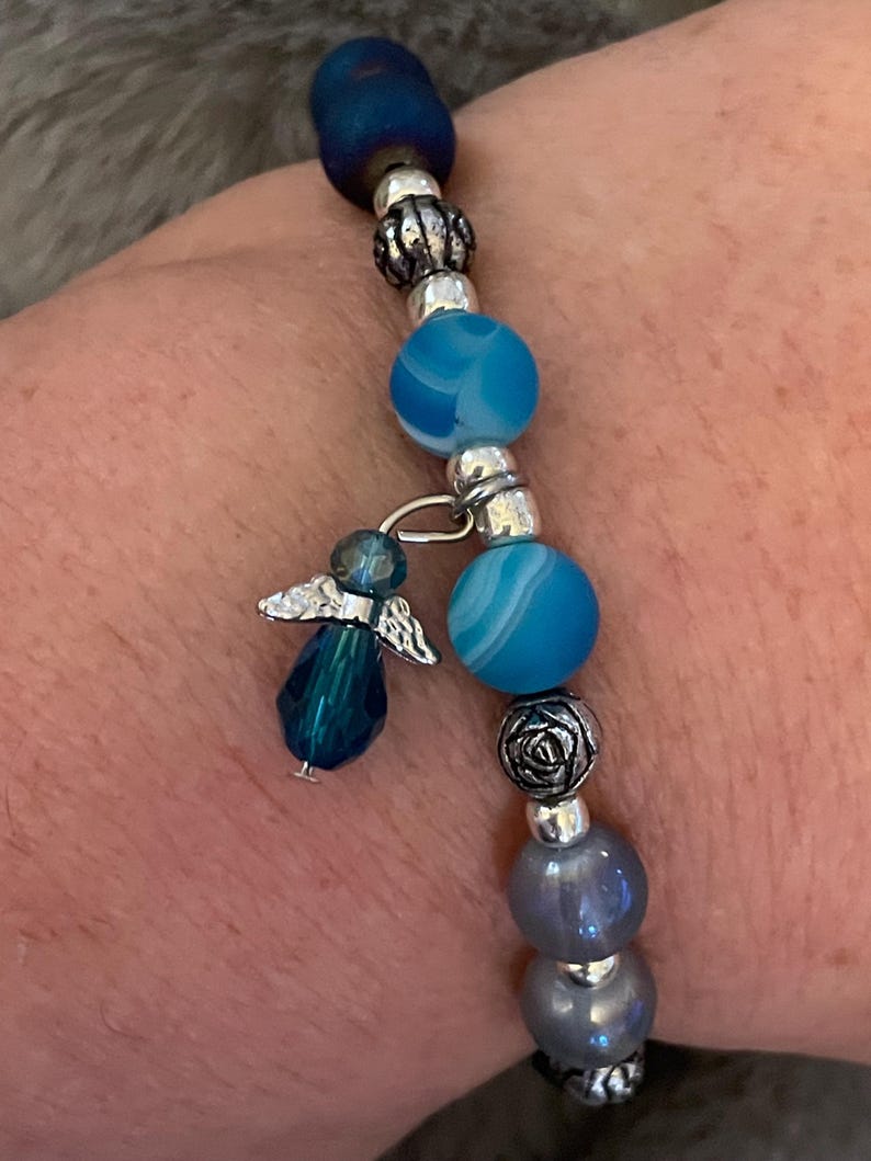 May include: A beaded bracelet featuring blue and silver beads, with a dangling angel charm. The bracelet includes various shades of blue beads, silver accents, and a small angel pendant. The bracelet is designed to be worn on the wrist.
