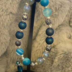 May include: A beaded bracelet featuring a mix of blue and iridescent beads, interspersed with silver-colored decorative beads. A small, dangling angel charm adds a delicate touch. The bracelet is displayed against a soft, gray background.