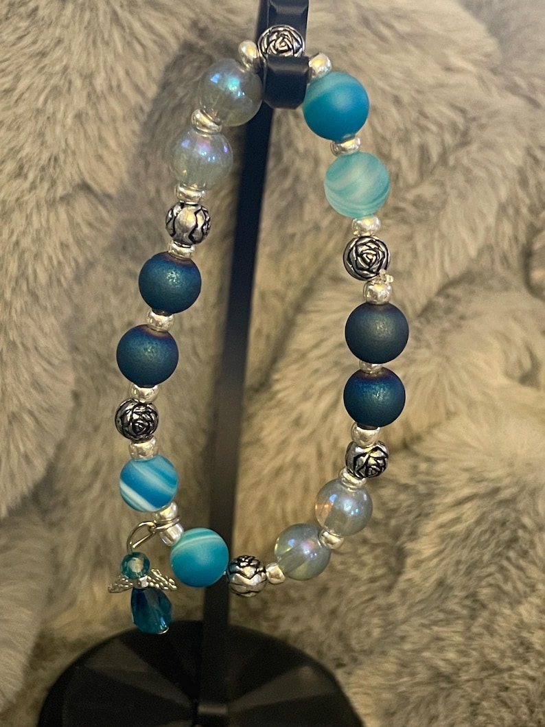 May include: A beaded bracelet featuring a mix of blue and iridescent beads, interspersed with silver-colored decorative beads. A small, dangling angel charm adds a delicate touch. The bracelet is displayed against a soft, gray background.