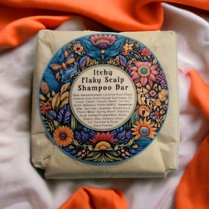 May include: A shampoo bar wrapped in beige paper, with a circular label displaying floral designs and the text "Itchy Flaky Scalp Shampoo Bar". The label lists ingredients. The bar is set against white and orange fabric.