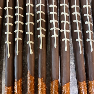 May include: A close-up of ten chocolate-covered pretzel rods with white icing and a football stitch design. The pretzels are arranged in a row on a gray surface.