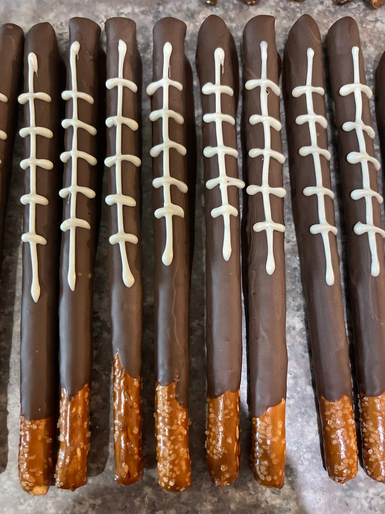 May include: A close-up of ten chocolate-covered pretzel rods with white icing and a football stitch design. The pretzels are arranged in a row on a gray surface.