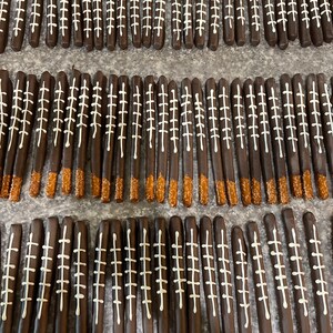 May include: A close-up of a tray of chocolate-covered pretzel rods decorated with white icing to resemble footballs. The pretzels are arranged in rows on a gray surface.