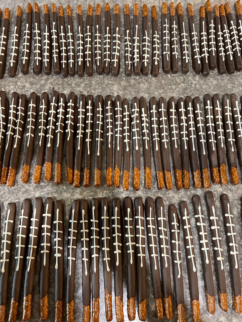 May include: A close-up of a tray of chocolate-covered pretzel rods decorated with white icing to resemble footballs. The pretzels are arranged in rows on a gray surface.