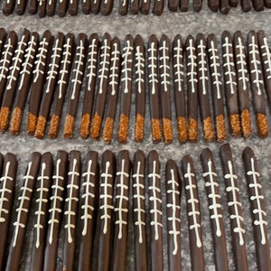 May include: A close-up of a large quantity of chocolate-covered pretzel rods decorated with white icing to resemble footballs. The pretzels are arranged in rows on a brown surface.