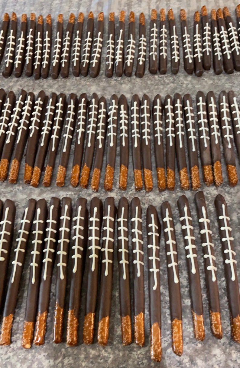 May include: A close-up of a large quantity of chocolate-covered pretzel rods decorated with white icing to resemble footballs. The pretzels are arranged in rows on a brown surface.