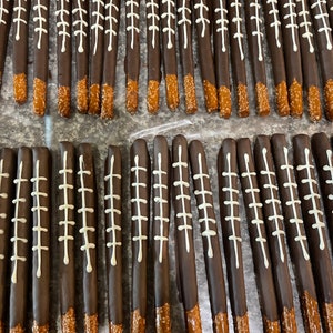 May include: A close-up of two rows of chocolate-covered pretzel rods with white football stitching designs. The pretzels are arranged in rows on a brown surface.