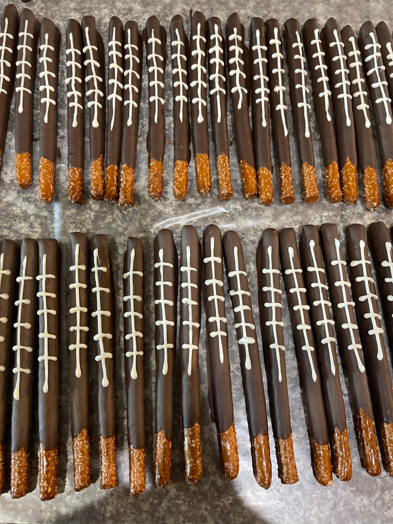 May include: A close-up of two rows of chocolate-covered pretzel rods with white football stitching designs. The pretzels are arranged in rows on a brown surface.