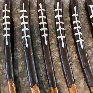 May include: A close-up of six chocolate-covered pretzel rods decorated with white icing to resemble footballs. The pretzels are arranged in a row on a brown surface.