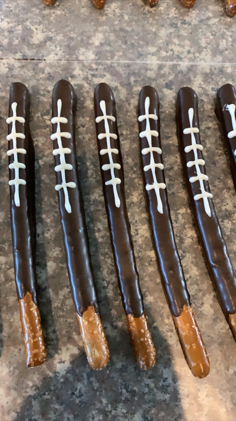 May include: A close-up of six chocolate-covered pretzel rods decorated with white icing to resemble footballs. The pretzels are arranged in a row on a brown surface.