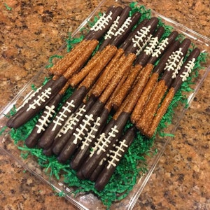 May include: A clear plastic tray filled with chocolate-covered pretzel sticks decorated with white icing to resemble footballs. The tray is sitting on a brown surface with green shredded paper.