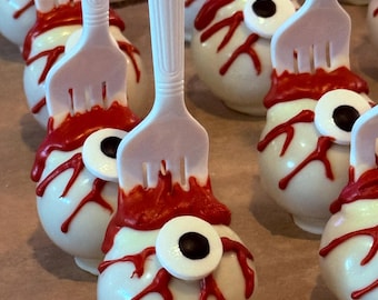May include: A collection of round cake pops shaped like eyeballs, each featuring a white coating, red icing veins, and a central iris with a black pupil. Each eyeball is pierced by a white plastic fork, creating a series of treats arranged in rows.