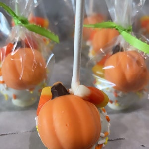 May include: A close-up of a pumpkin-shaped cake pop decorated with white and yellow sprinkles. The cake pop is on a stick and is topped with a candy corn and a chocolate piece. The cake pop is wrapped in clear cellophane and tied with a green ribbon.