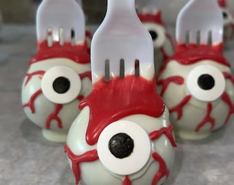 Puede incluir: Cuatro cake pops de ojo de chocolate blanco con glaseado rojo y pupilas negras en tenedores blancos.