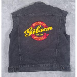 May include: A black denim vest featuring a large embroidered patch on the back. The patch displays the yellow script text Gibson, with red banners above and below reading Custom Art and Historic, and the text Since 1894 in small yellow letters.