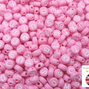 以下が含まれることがあります： 白い文字が書かれたピンクのプラスチック製のアルファベットビーズがたくさんあります。ビーズは丸く、中央に糸を通すための穴があります。