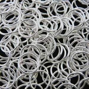 May include: A close-up shot of numerous silver jump rings. The rings are circular and have a shiny, metallic appearance. They are arranged densely against a dark background, forming a textured pattern. Suitable for jewellery making and crafts.