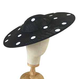 May include: A black hat with a wide brim and a smaller, rounded crown. The hat is adorned with white polka dots. The hat is displayed on a mannequin head with a wooden stand. The hat is a fashion accessory.