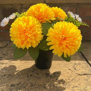 May include: A floral arrangement featuring vibrant yellow, spherical chrysanthemum-like flowers and small white daisy-like blossoms with green leaves. The flowers are contained within a simple, solid black cylindrical pot.