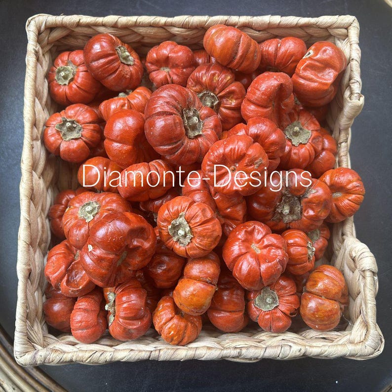 May include: A square woven basket filled with numerous small, round, orange pumpkins with visible stems. The pumpkins have a ribbed texture and a glossy finish. The text "Diamonte-Designs" is overlaid in white across the center of the image.