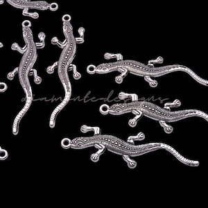 Puede incluir: Encantos de lagarto plateados con escamas detalladas y un lazo para sujetar a joyas o manualidades. Los encantos están dispuestos sobre un fondo negro, con el texto "diamante-designs" visible. Los encantos miden aproximadamente 5 cm de largo.