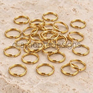 May include: A collection of gold-coloured metal jump rings, each featuring a circular shape with a split opening. The text "diamontedesigns" is overlaid across the centre of the pile of metallic rings, which are arranged in a loose, scattered cluster.