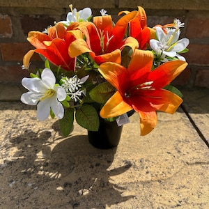 Könnte beinhalten: Ein lebendiger Strauß künstlicher Lilien in einem schwarzen Topf. Das Arrangement zeigt große orange und gelbe Lilien, durchsetzt mit kleineren weißen Blüten und grünen Blättern. Die Blumen stehen vor einer Backsteinmauer und einer Steinoberfläche.