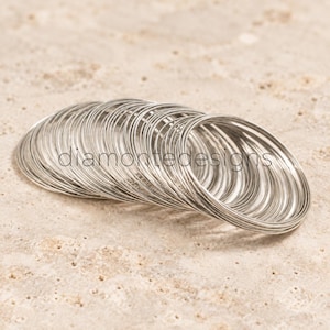 Puede incluir: Una pila de anillos metálicos circulares y delgados de tono plateado dispuestos en una formación ligeramente superpuesta e inclinada. La palabra diamontedesigns está impresa en una fuente sans-serif oscura y minúscula.