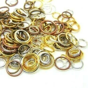 Op de afbeelding: Een verzameling metalen verbindingsringen in goud, zilver en koper. De ringen variëren in grootte en zijn verspreid over een witte ondergrond. Ideaal voor het maken van sieraden en knutselprojecten.