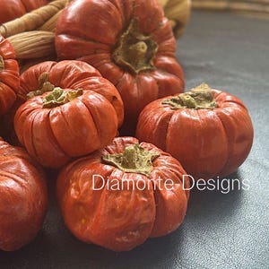 May include: A cluster of small, round, orange-red pumpkins with deep vertical ridges and brown stems. The pumpkins have a textured, slightly wrinkled surface. The text Diamonte-Designs is printed in white across the lower portion of the image.