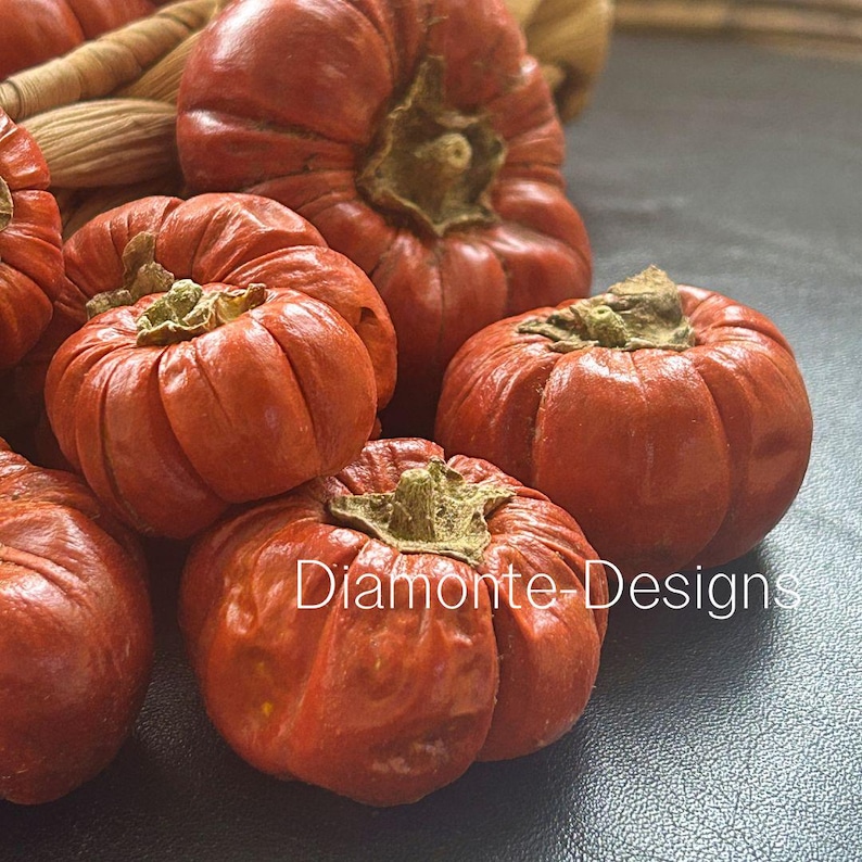 May include: A cluster of small, round, orange-red pumpkins with deep vertical ridges and brown stems. The pumpkins have a textured, slightly wrinkled surface. The text Diamonte-Designs is printed in white across the lower portion of the image.